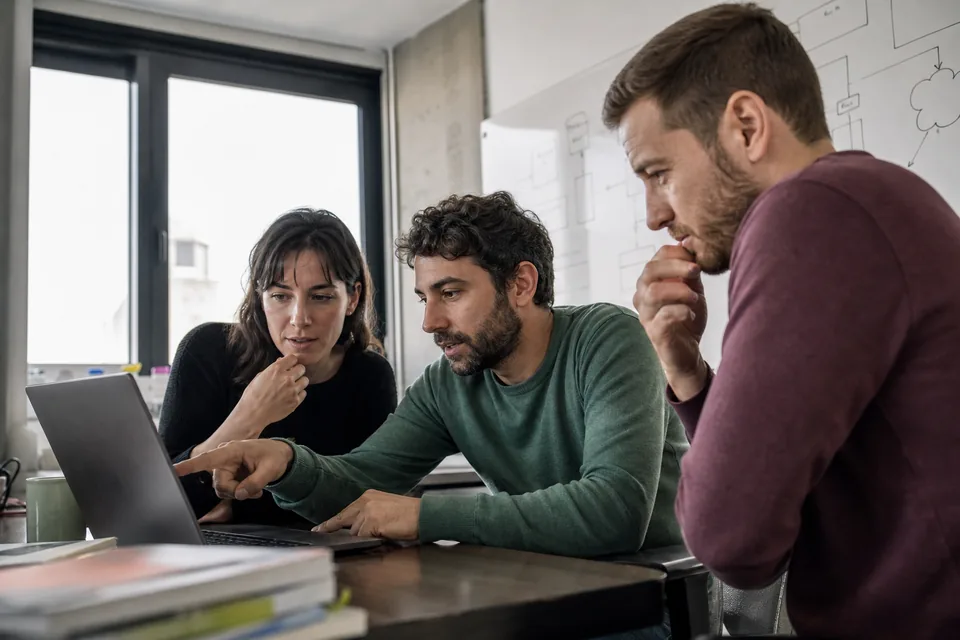 Three developers reviewing a monthly usage bill on a laptop next to an architecture whiteboard