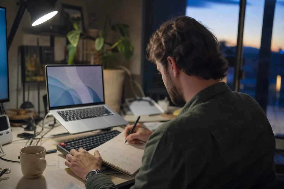 Developer at a home desk searching a large codebase on a laptop while taking notes