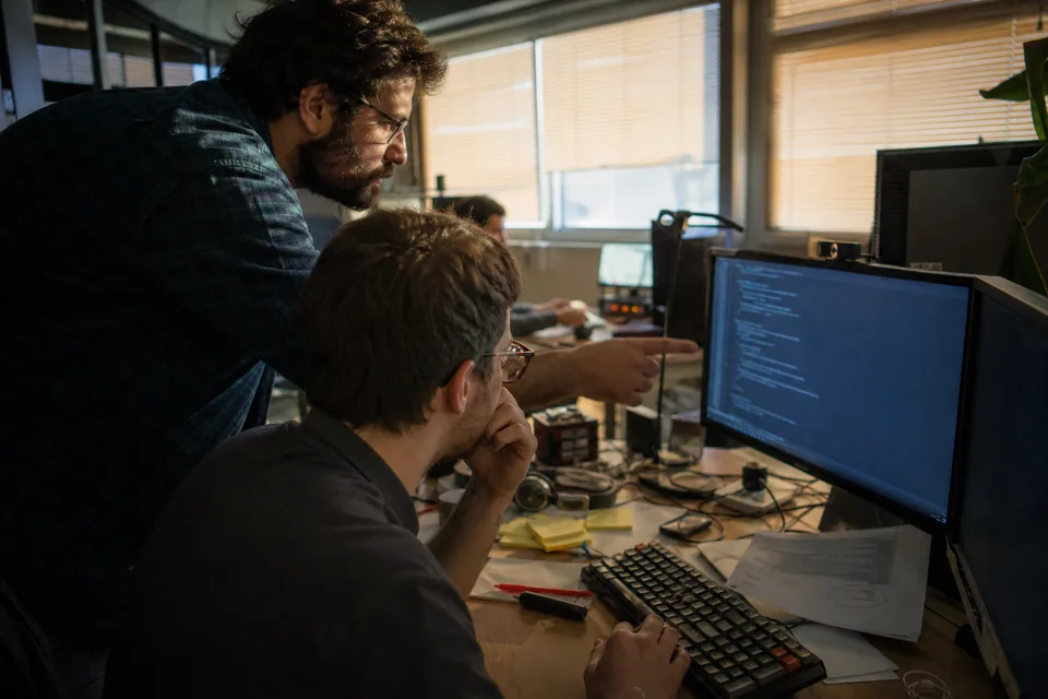Two developers at a cluttered desk reviewing code next to a printed invoice with circled totals