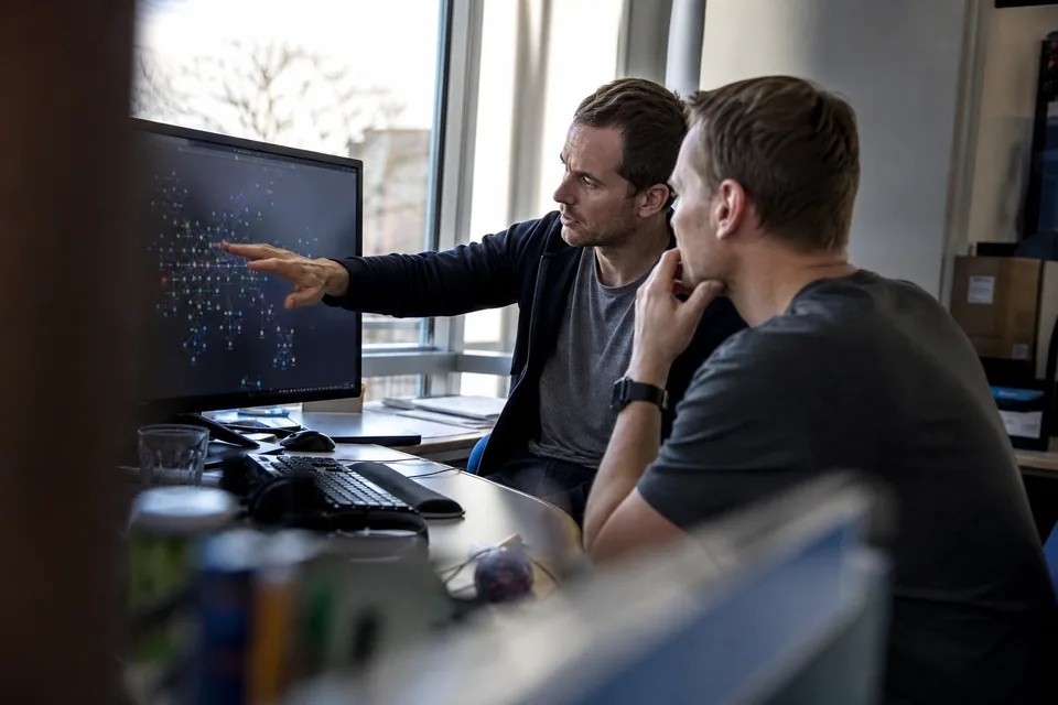 Two software engineers discussing a dependency graph on a large monitor in an office